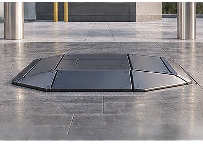 The pedestal mount fully retracted: a faceted lid sitting flush in the paving where the mount stands, with the surrounding slabs and bollards of the plaza around it.