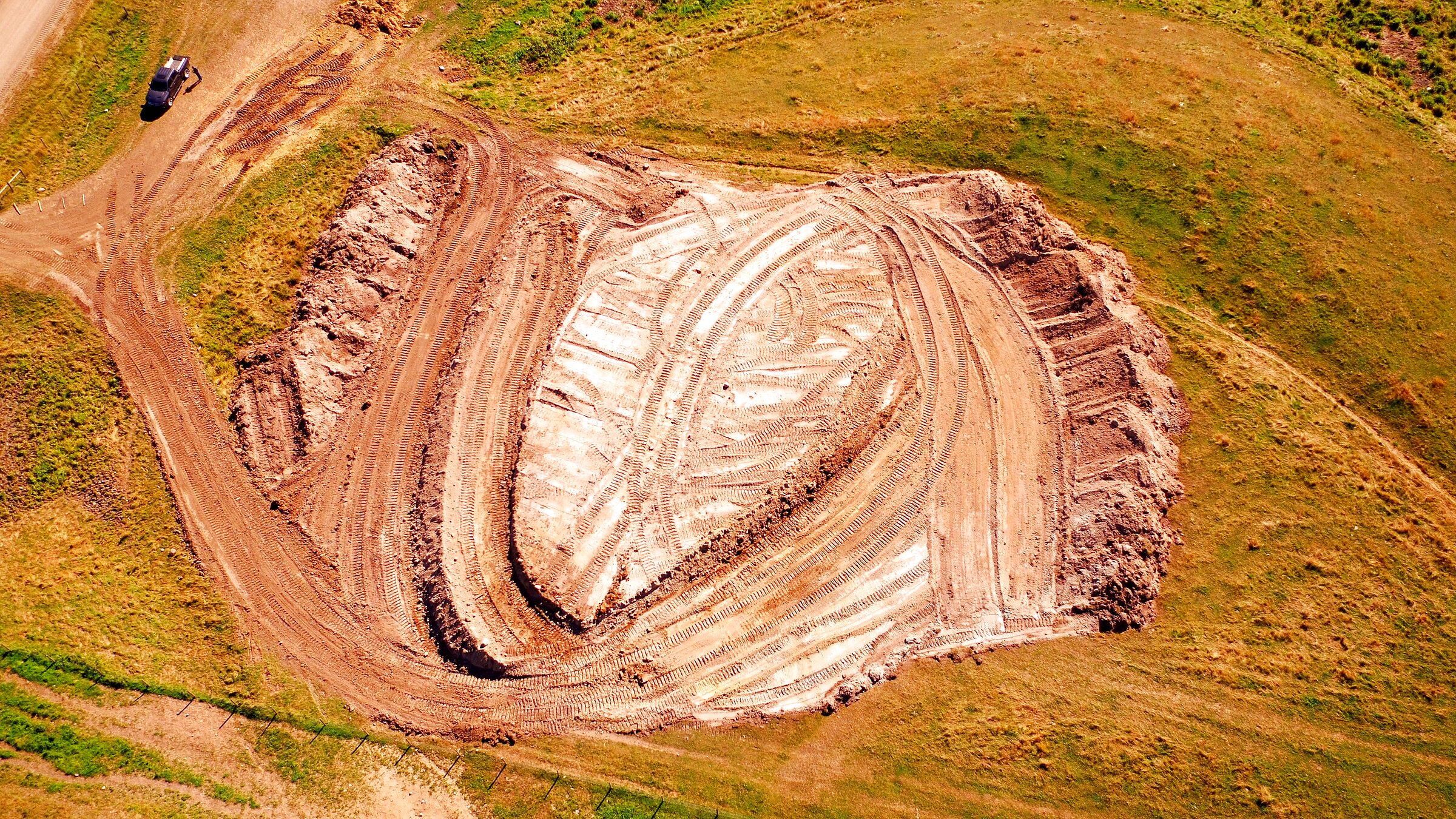 A borrow pit seen from the air: the cut face, the haul tracks curving through the open ground, and a pickup parked at the edge of the grass.