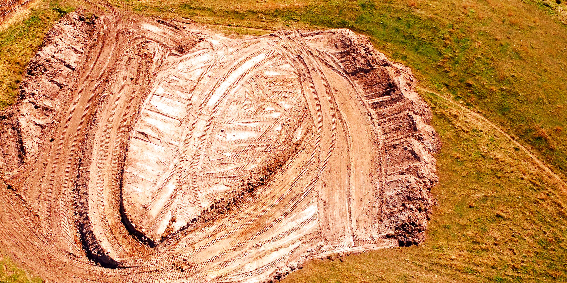 A borrow pit seen straight down from the air: the cut floor, the benches and ramps around it and the overlapping passes of the machines that cut it, on open grassland.