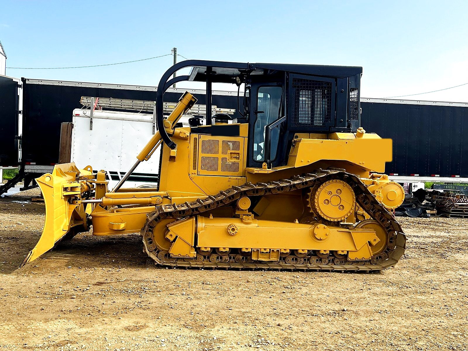 The group's own D6T LGP dozer in side profile on a gravel yard: the blade lowered at the front, the screened cab and its guard above the track frame, and parked trailers behind it.