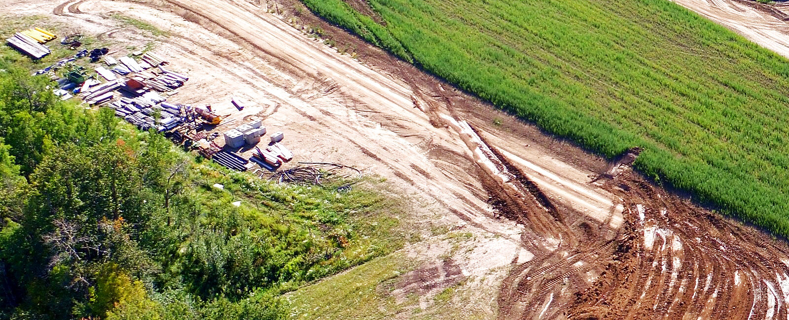 A laydown yard from the air: pipe, timber and material stacked in rows on bare ground beside a cut running away between a treeline and a standing crop, with plant parked at the far end of the spread.