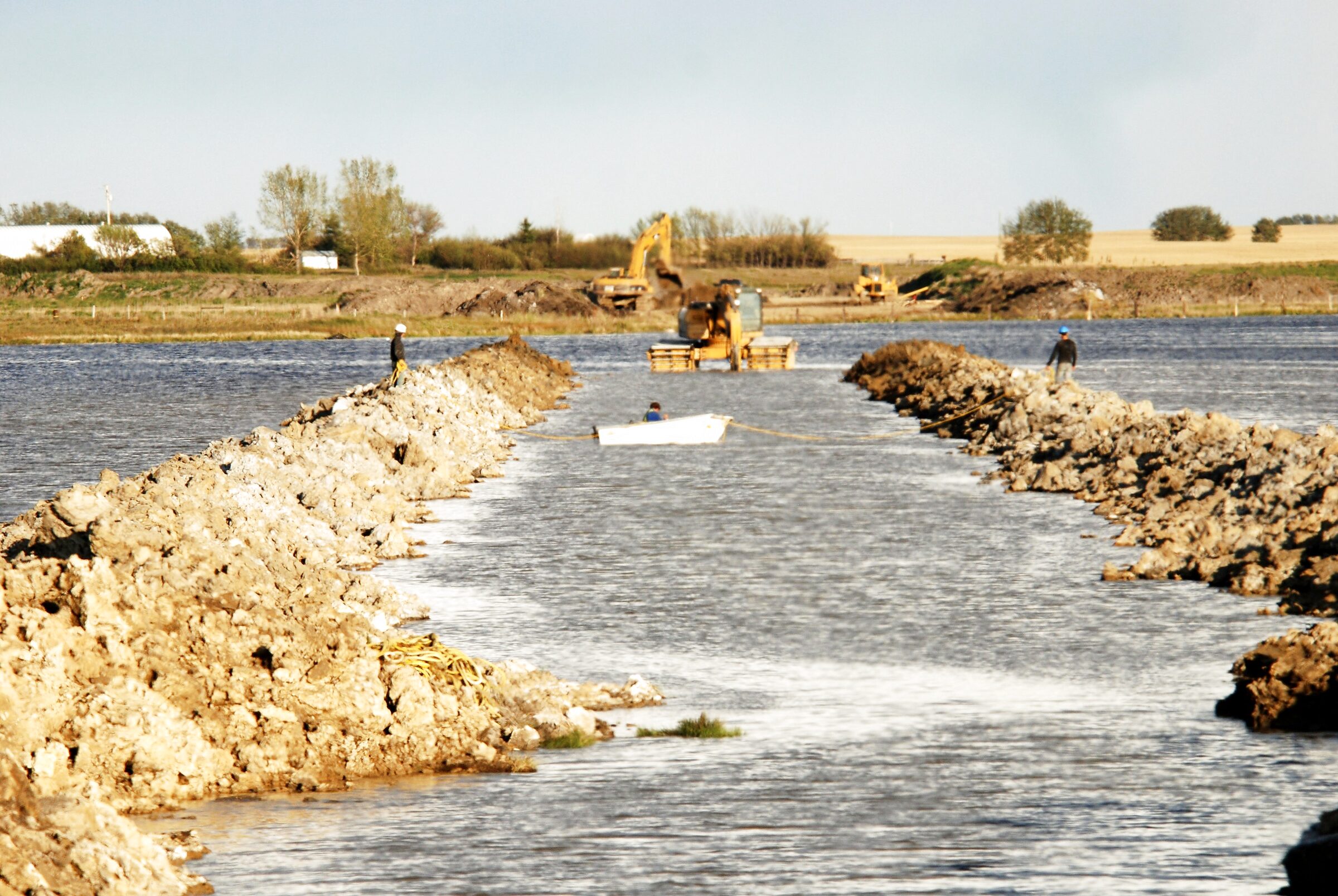 Two of the group's crew on the arms of a rock causeway being closed across open water at Weed Lake, Alberta, an excavator on pontoons and a small boat on the channel between them.