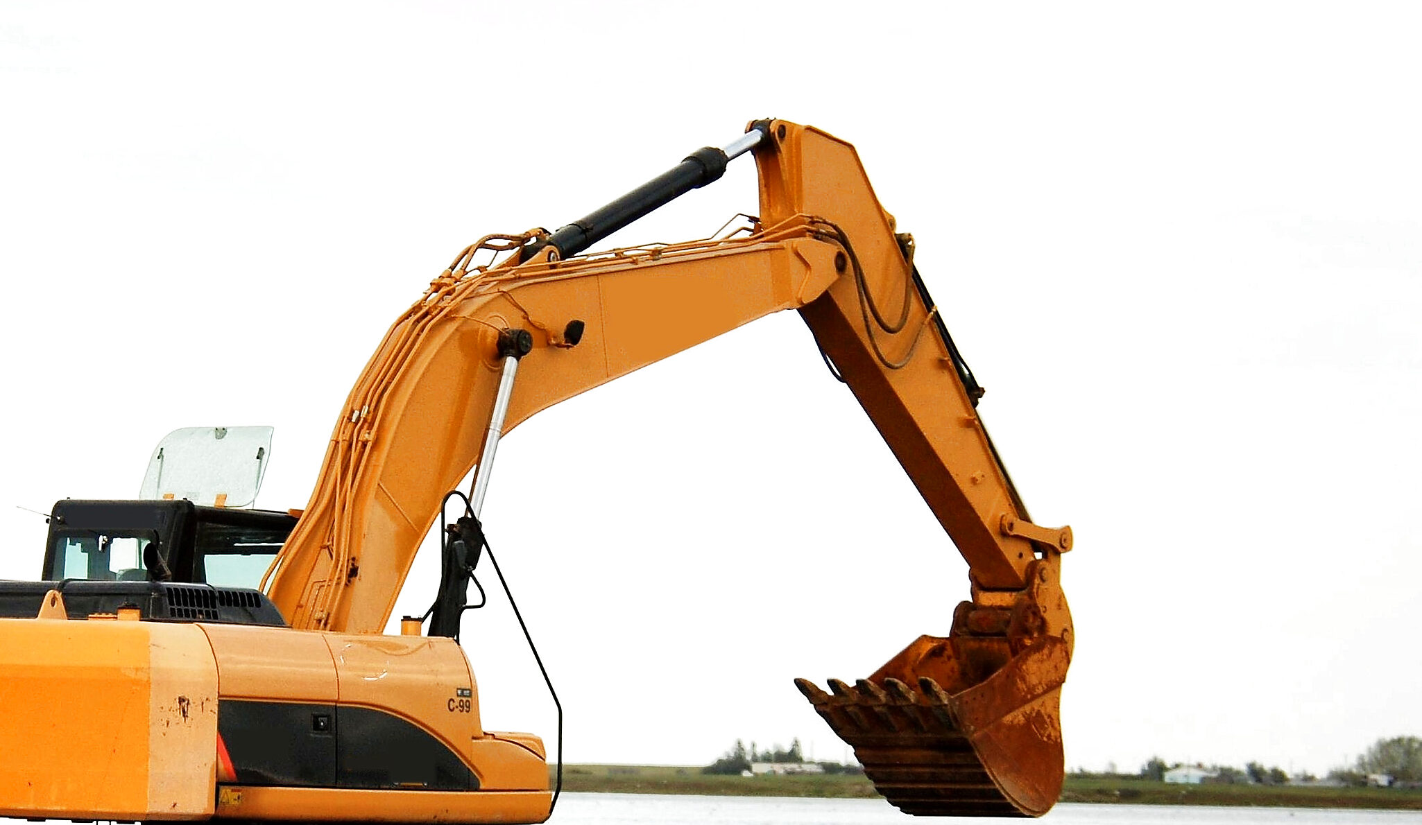 The boom, stick and bucket of an excavator raised against a bright sky, the hydraulic cylinders and hoses that work them running along the boom, the cab below.