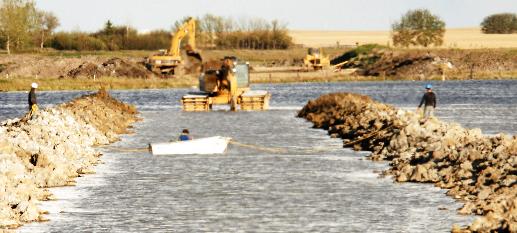 Two of the group's crew standing on the rock arms of a causeway being closed across open water, an amphibious excavator on pontoons working the channel between them and a small boat on the water below it.