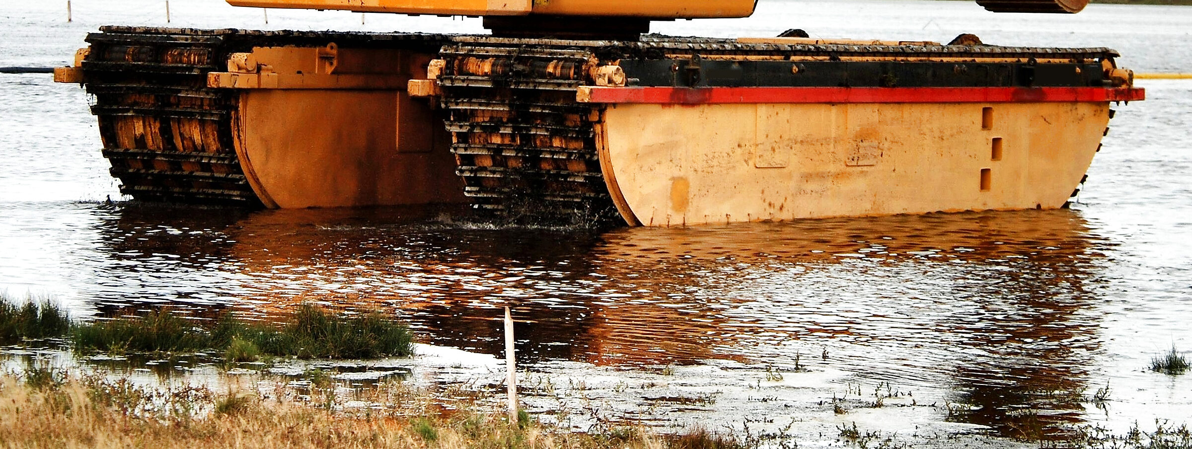 The undercarriage of an amphibious excavator: two steel tracks running over long pontoon floats, mud on the track plates and water lapping at the hulls.