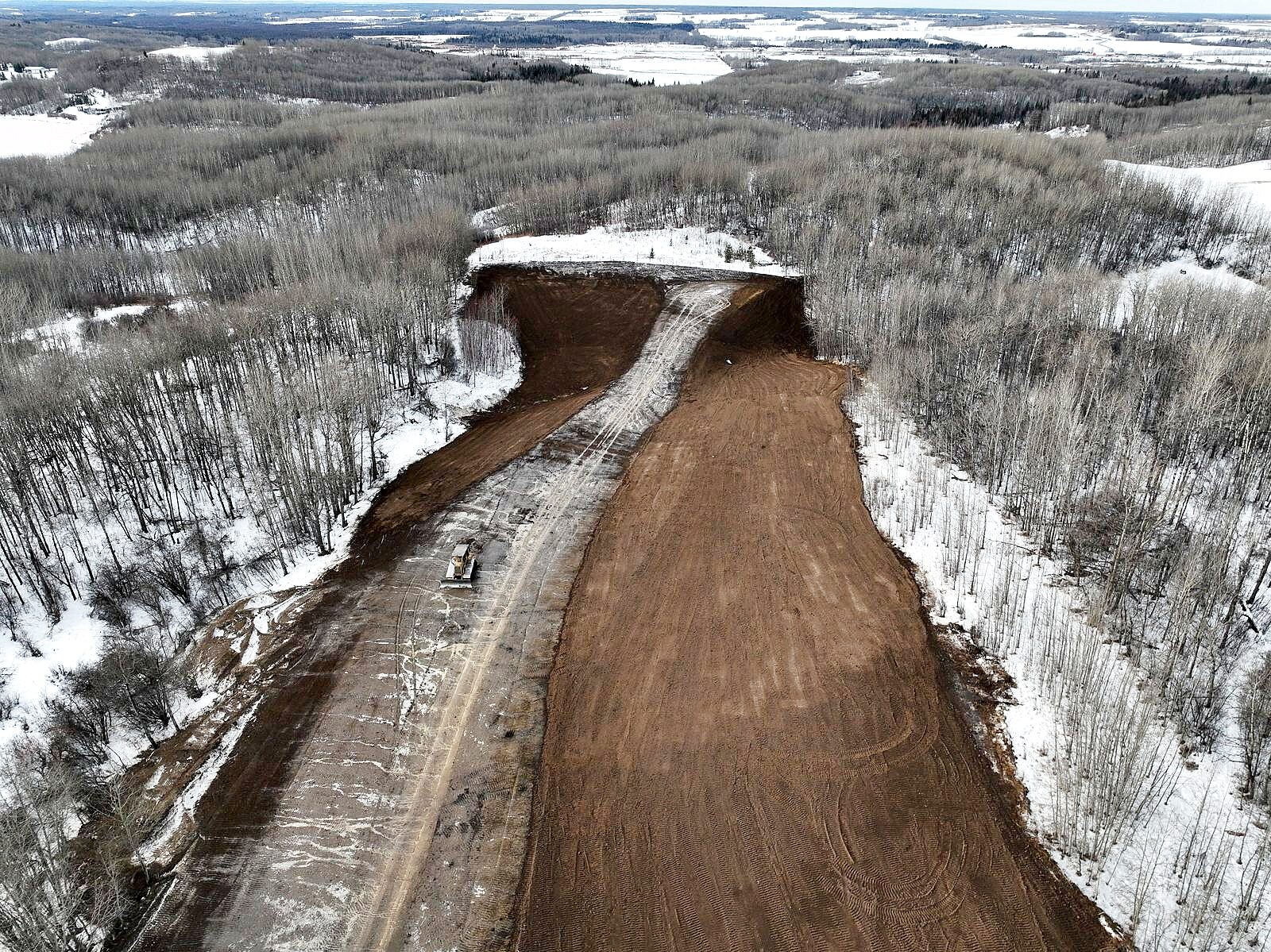 A dozer on a freshly cut corridor through winter forest, seen from the air, the graded ground dark against the snow.